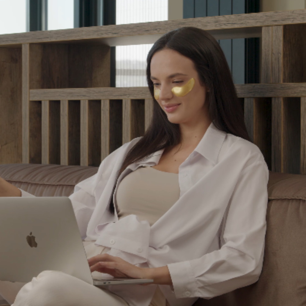 A woman relaxing on a bed wearing 24k gold eye mask under her eyes.